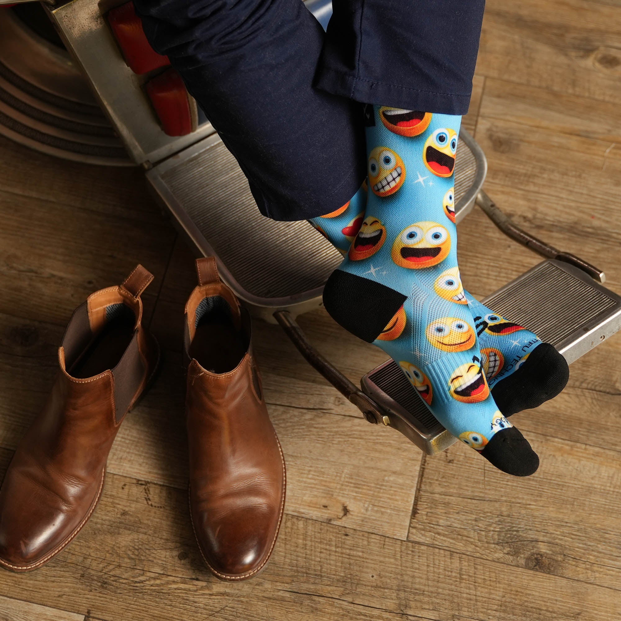 Person sitting in  barber's chair with feet up wearing blue trousers and fun emoji Sox Footwear. The socks have a light blue background with bright yellow smiley faces on them.
